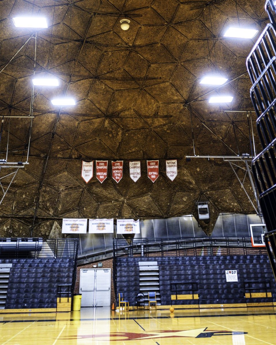Wide view from the baseline inside the Dome gym showing the hexagon-patterned brown ceiling, bright overhead lights, folded black bleachers, and red and white championship banners hanging above the court.