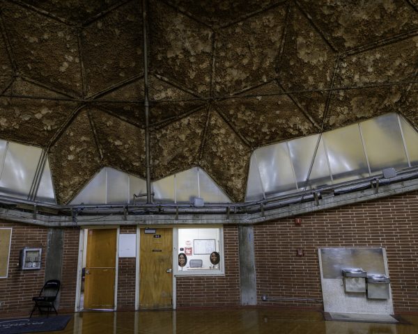 Panoramic view of the Dome’s interior ring, showing brick walls, wooden doors, a small office window and water fountains under a jagged band of translucent panels and rough brown ceiling.