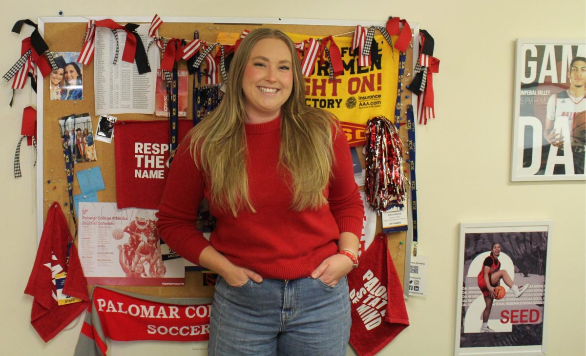 Cara Heise, wearing a red sweater stands in front of a wall decorated with various sports photos and flags.