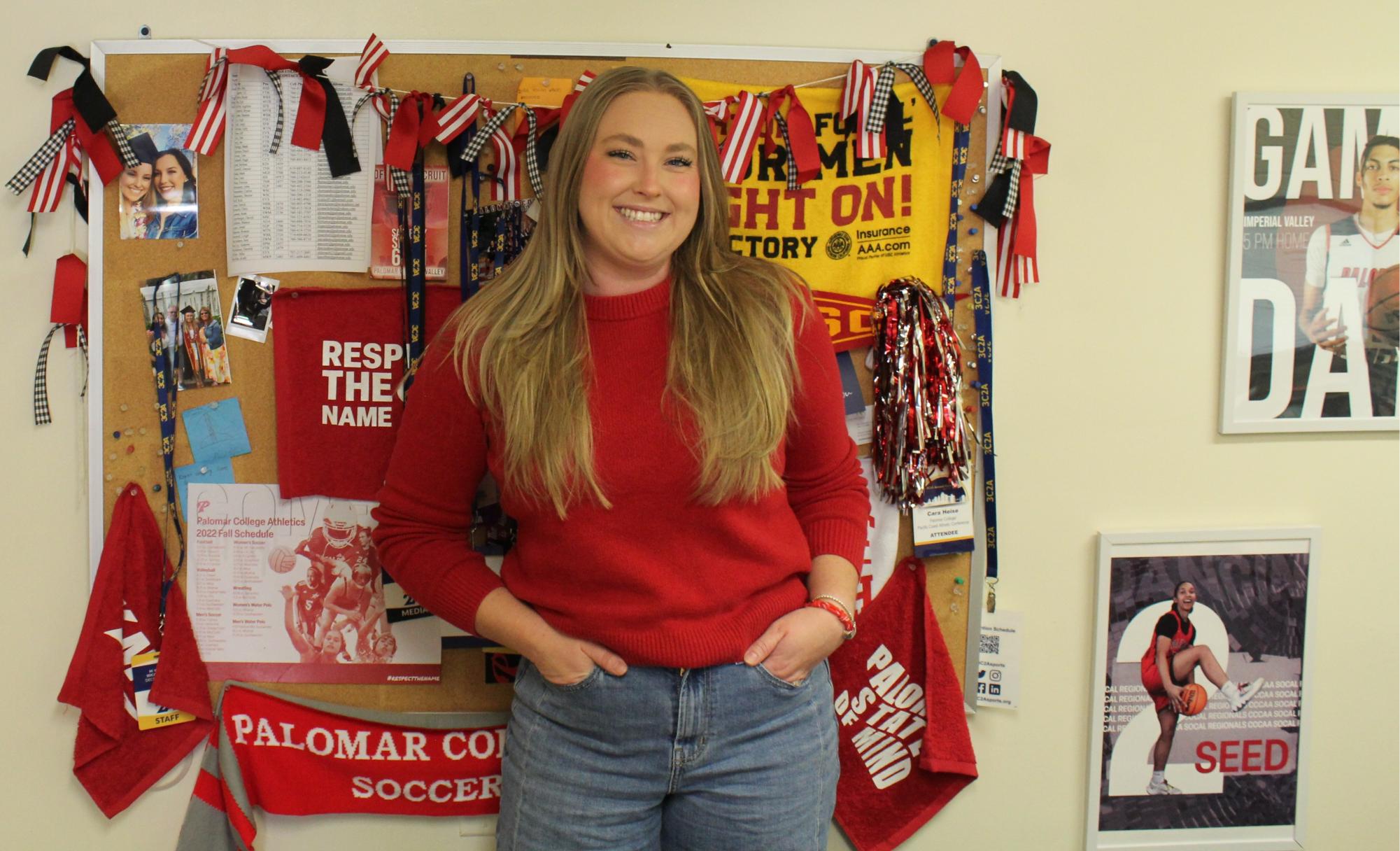 Cara Heise, wearing a red sweater stands in front of a wall decorated with various sports photos and flags.