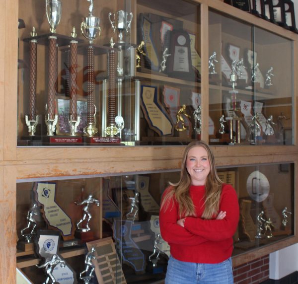Cara Heise, wearing a red sweater stands in front of a wooden cabinet filled with various trophies won by Palomar College's athletic programs.