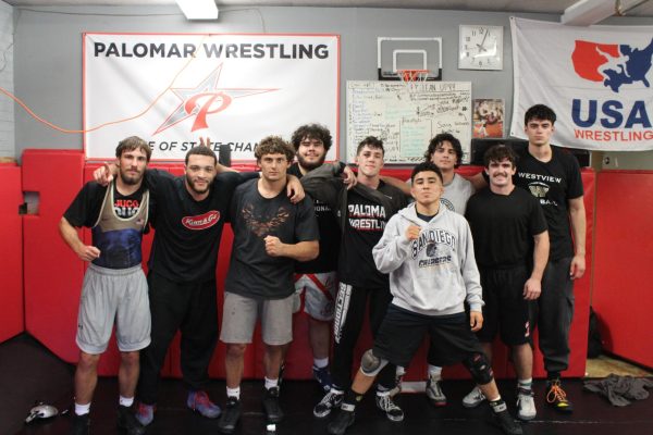 Nine members of the Palomar men's wrestling team stand side by side in casual workout clothes in the wrestling room at Palomar College on December 10, 2025.