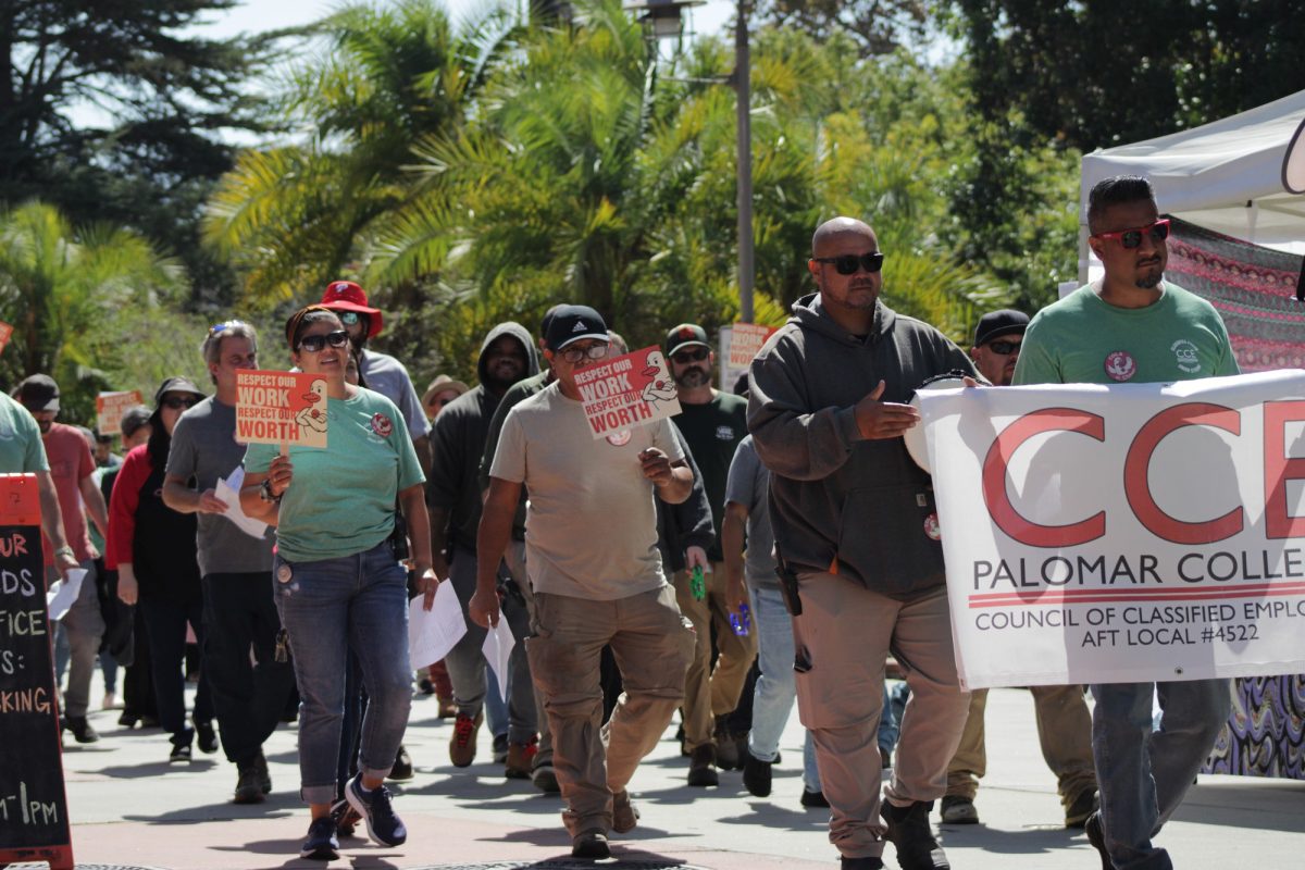 A group of people walk down a campus pathway, many wearing green shirts and carrying orange “Respect our work, respect our worth” signs, while two members at the front hold a large white banner that reads “CCE Palomar College Council of Classified Employees.”