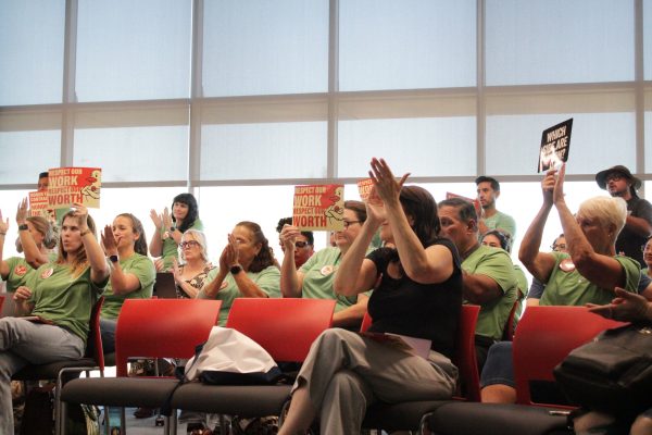 A row of classified employees wearing green shirts sit in red chairs, clapping and holding orange signs that read “Respect our work, respect our worth” during a public meeting.