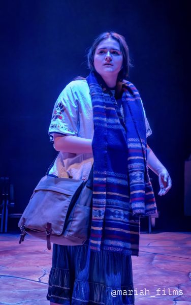 Joven con blusa bordada, falda larga, bolso cruzado y bufanda azul a rayas mira hacia arriba con expresión preocupada bajo una luz azul fría.