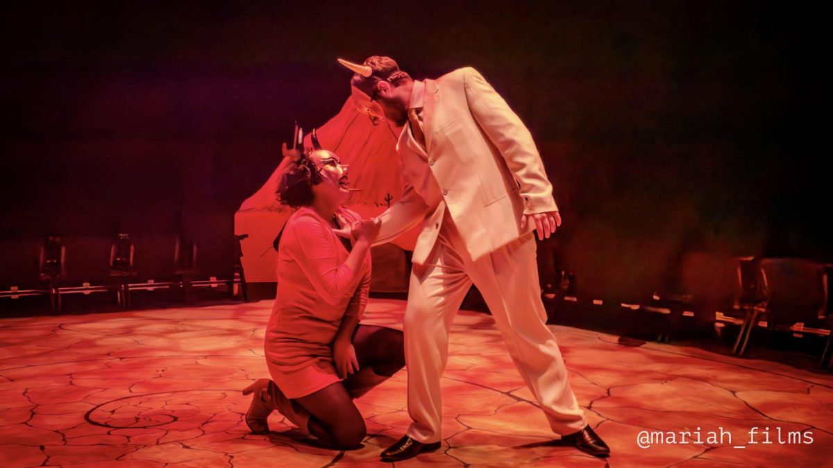 Un intérprete con traje blanco y máscara con cuernos se inclina sobre un demonio arrodillado con vestido rojo; se toman de las manos en un momento intenso sobre un escenario iluminado de rojo, rodeado por el público del estudio.