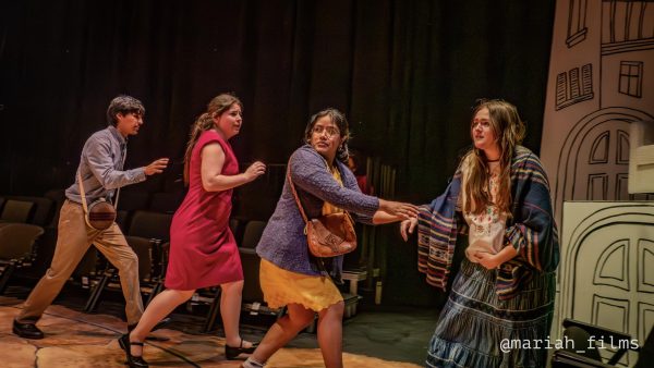 Four student actors run across a circular stage: a young man with a round canteen bag, a woman in a red dress, a woman in a yellow dress and purple cardigan reaching back, and a pregnant woman in a long skirt and striped shawl, all playing shepherds in "Pastorela Fronteriza."