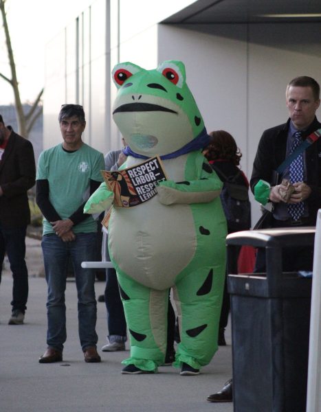 Matt Estes is in a frog inflatable suit holding a sign that says "respect labor, respect learning" and is standing outside the library.