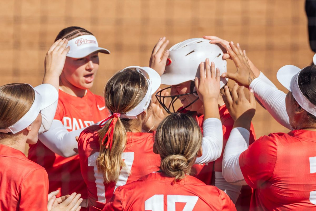 Palomar softball player Alexis Huey in a white helmet and red jersey celebrates her leadoff home run with her teammates touching her helmet at home plate.