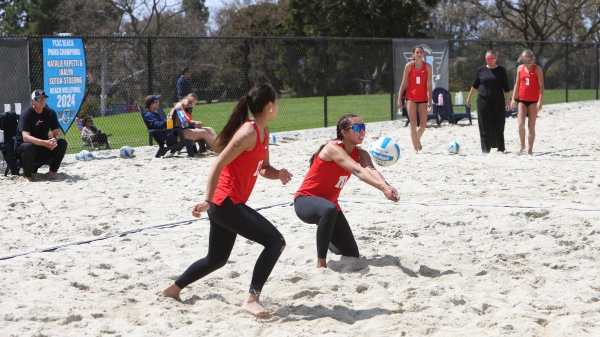 A beach volleyball player in a red jersey and sunglasses extends her arms to pass an incoming volleyball while her teammate starts running toward the volleyball net.