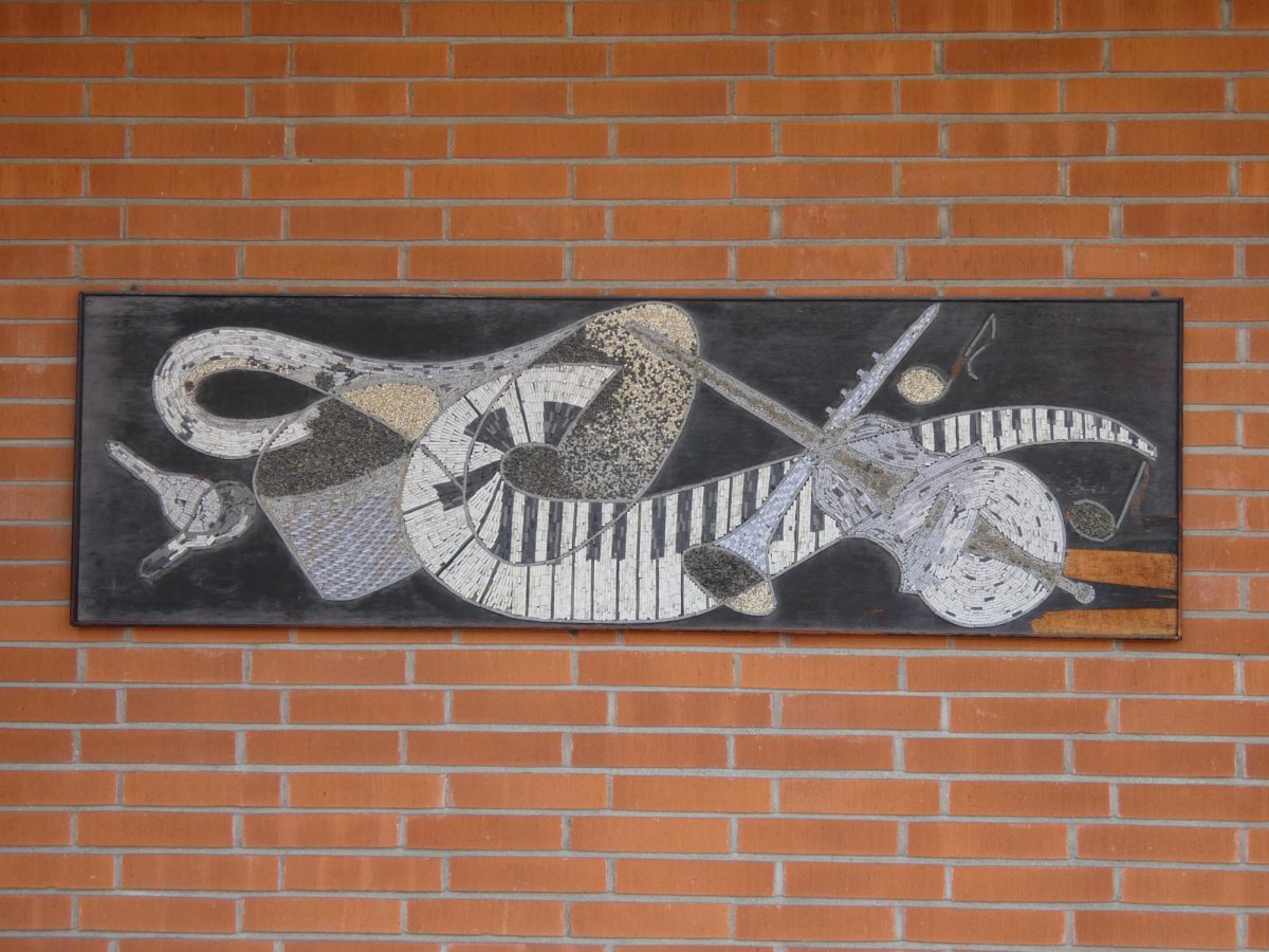 A mosaic mural on a brick wall, made of small white, gray, and beige tiles arranged in sweeping, abstract shapes. Piano keys and string-instrument forms curve across a dark background in a flowing, music-inspired design.