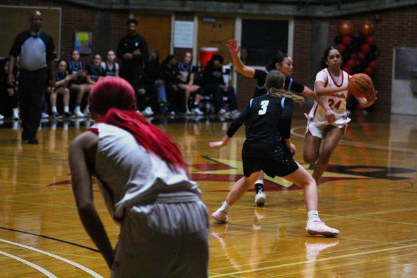 Palomar basketball player Kylee Trujillo in a white uniform with red hair waits at her own 3-point line as her teammate Ivorii Seals, number 24 dribbles around two MiraCosta defenders in black uniforms on Feb. 20 at the Dome.