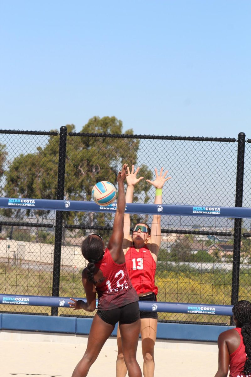 Palomar beach volleyball player Reese Maffitt (#13) in red leaps and reaches to block a hit from Mt San Jacinto's #12 in red/black.