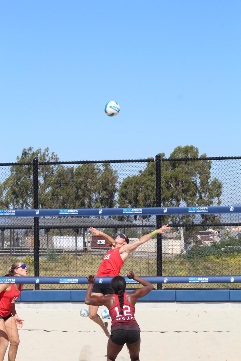 Palomar beach volleyball player Reese Maffit (#13) in red leaps with her arms outstretched and prepares to spike a volleyball.