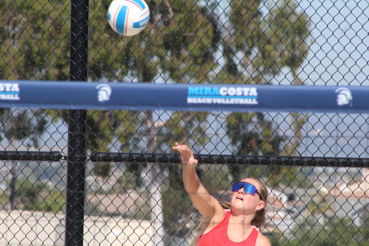 Palomar Comets beach volleyball player in red swings her right arm to serve.
