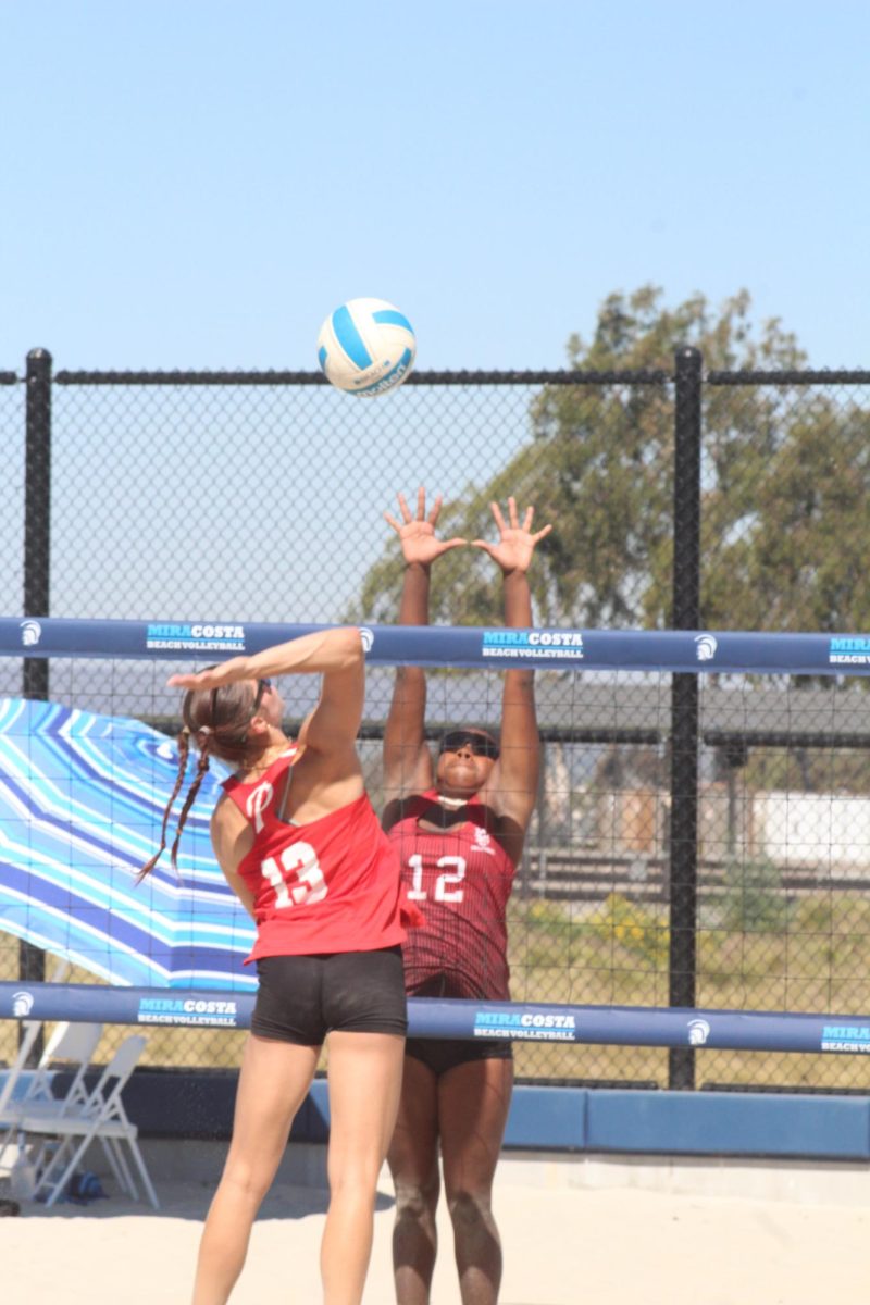 Palomar beach volleyball player Reese Maffitt (#13) in red prepares to spike the ball at a Mt San Jacinto player in red/black jumping for a block