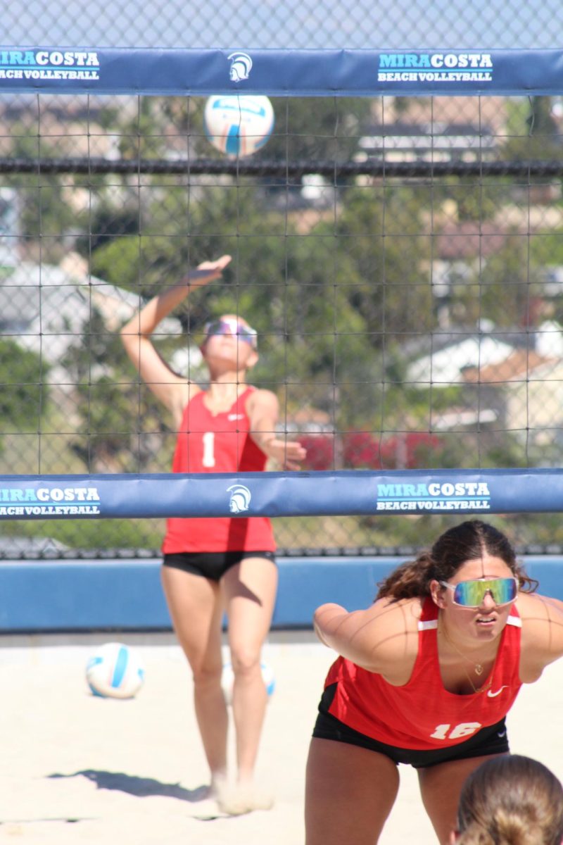 Palomar beach volleyball players in red jerseys, Grayson Grein (#1) throws the volleyball up to serve while her teammate Denise Garcia (#16) looks towards their opponents.