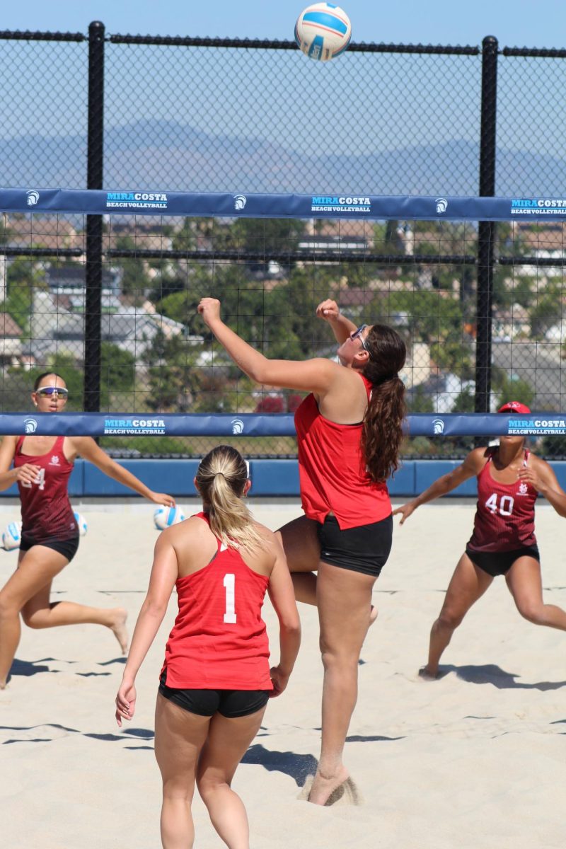 Palomar beach volleyball player Grayson Grein (#1) watches her teammate Denise Garcia, both in red, jump to hit the volleyball over the net at her opponents.