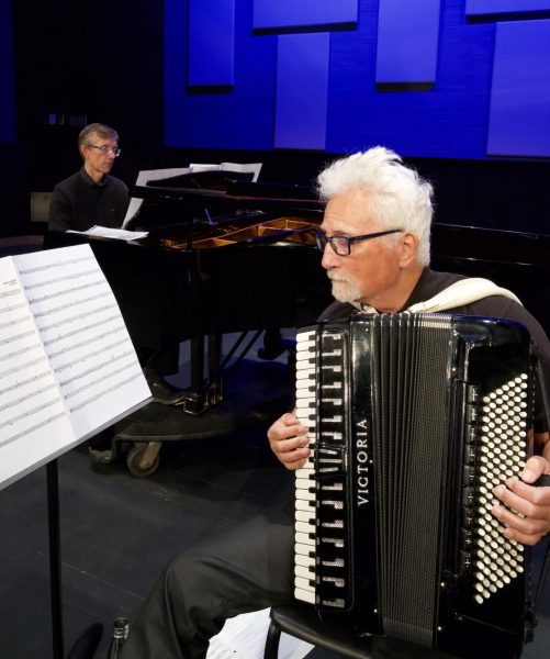 An accordionist plays in the foreground while a pianist accompanies on a grand piano in the background under blue stage lighting.