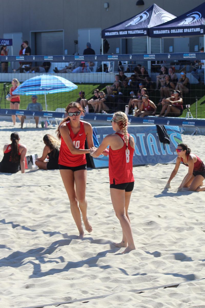 Palomar beach volleyball players in red jerseys, slap hands in front of the net after a successful point.