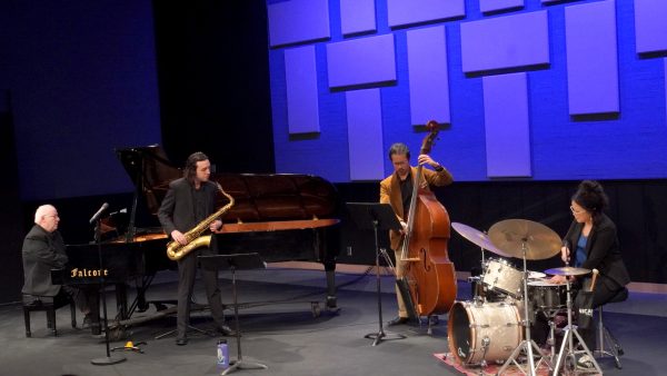 A jazz quartet performs onstage with piano, saxophone, upright bass and drums under blue lighting.