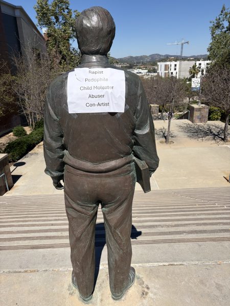 Estatua de bronce de Cesar Chavez vista desde atrás en la cima de una larga escalera al aire libre. Un cartel que dice "Violador, pedófilo, abusador de niños, maltratador, estafador" en negro sobre papel blanco está pegado en la parte trasera de la estatua.