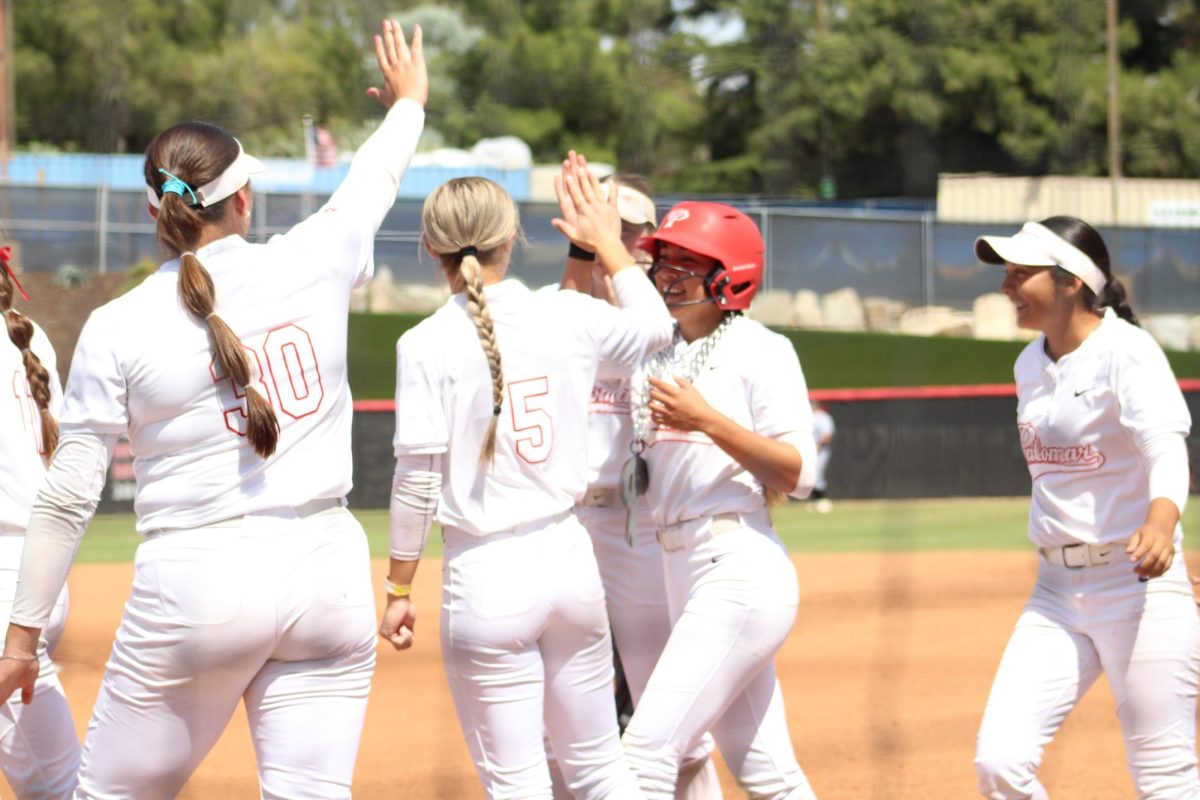 Palomar softball player Gisele Gonzalez wearing a red helmet and a white uniform celebrates with high fives for her teammates at home plate after hitting a solo home run.
