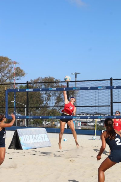 A beach volleyball player in red jumps to spike the ball while being close to the net.
