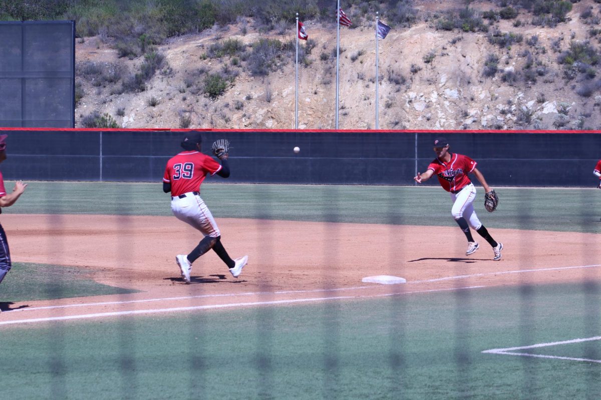Palomar first baseman in red tosses the baseball underhand to another Palomar player as he approaches first base to get the out at first.