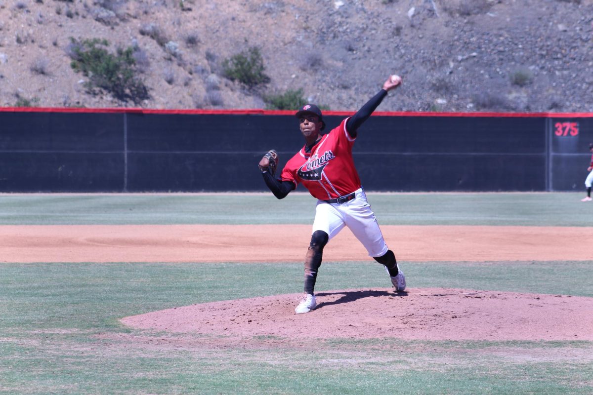 Palomar baseball pitcher in a red uniform has his left arm outstretched with the ball in his hand before releasing a warm-up pitch.