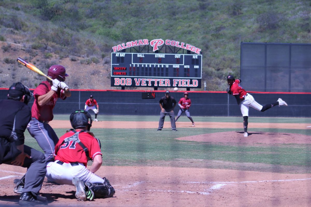 Palomar baseball pitcher in red pitches a ball that is halfway to home against a SD City batter in maroon.