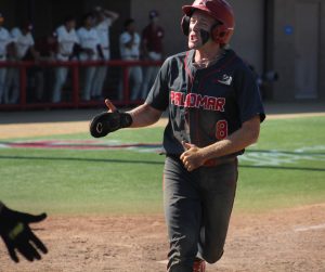 A Palomar baseball player in a dark grey uniform reaches his hand out for a high-five from his teammate after scoring a run.