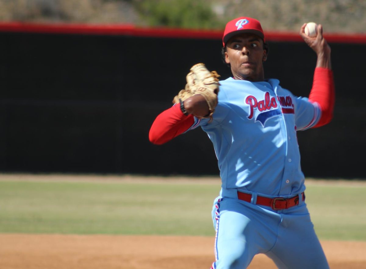 A Palomar College baseball pitcher in a light blue uniform reaches his left arm back mid pitch.