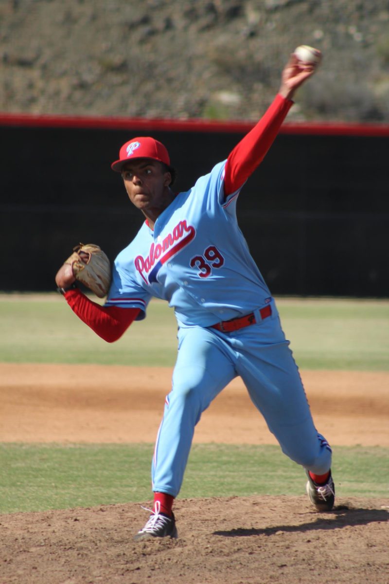 Palomar College baseball pitcher in a light blue uniform is mid pitch, striding towards the mound with the ball still held over his head.