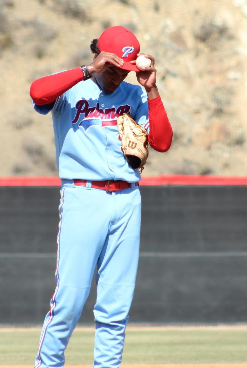 Palomar baseball pitcher in a light blue uniform adjusts his red hat with both hands, a baseball is held between his left hand and the brim of the hat.