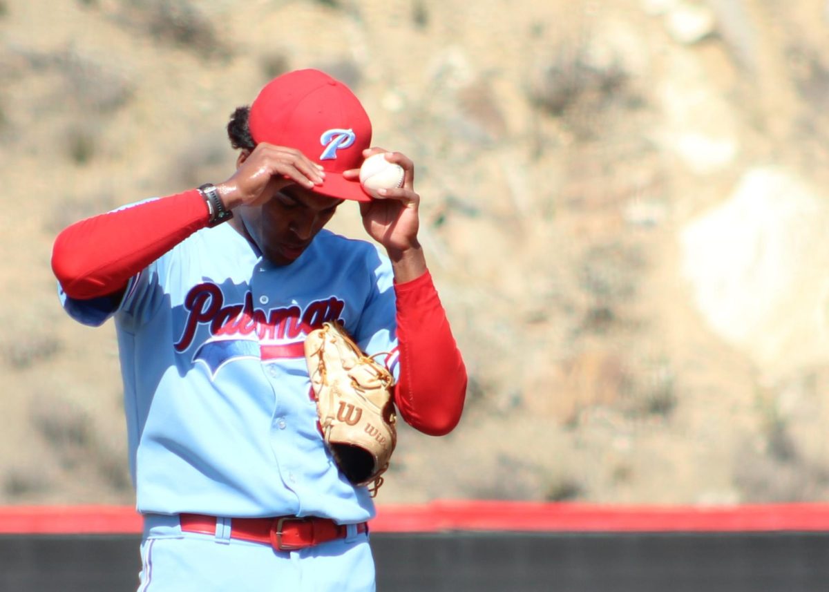 Palomar baseball pitcher in a light blue uniform adjusts his red hat with both hands, a baseball is held between his left hand and the brim of the hat.