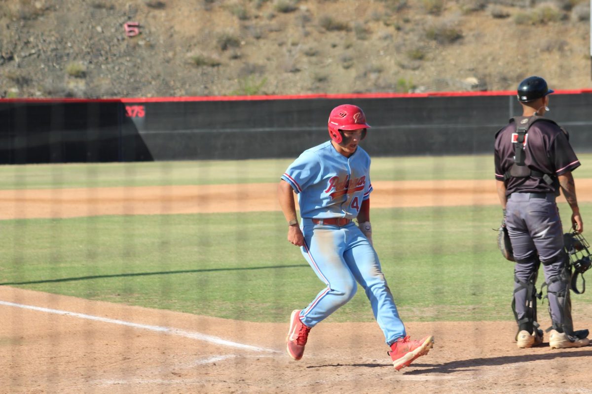 Palomar baseball player in a light blue uniform is about to touch home plate with his left foot while a Mt. San Jacinto catcher looks out toward the right side of the field.