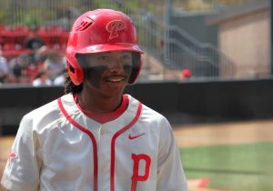 A palomar baseball player in a white jersey with red trim and a red helmet stares to the right in the on-deck circle.