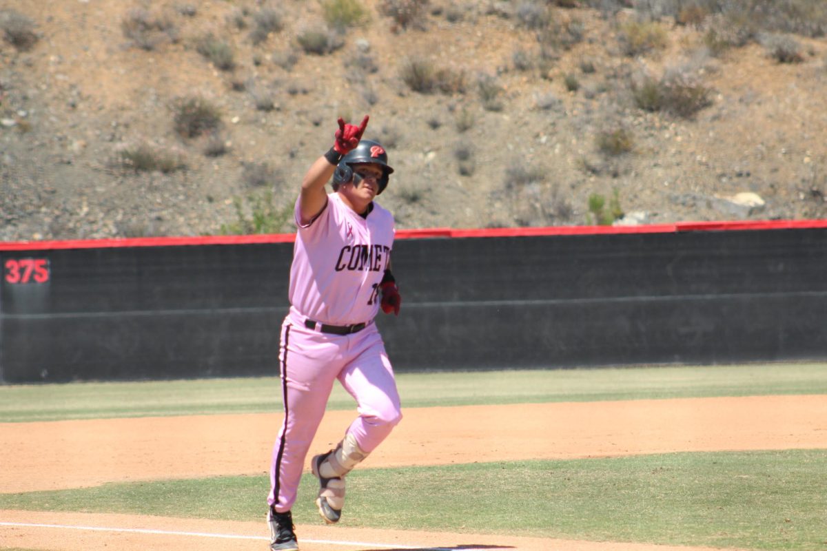 Palomar baseball player in a pink uniform raises his right hand in the shape of horns as he runs toward home plate following a three-run home run.