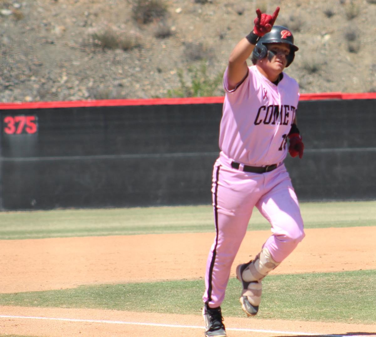 Palomar baseball player in a pink uniform raises his right hand in the shape of horns as he runs toward home plate following a three-run home run.